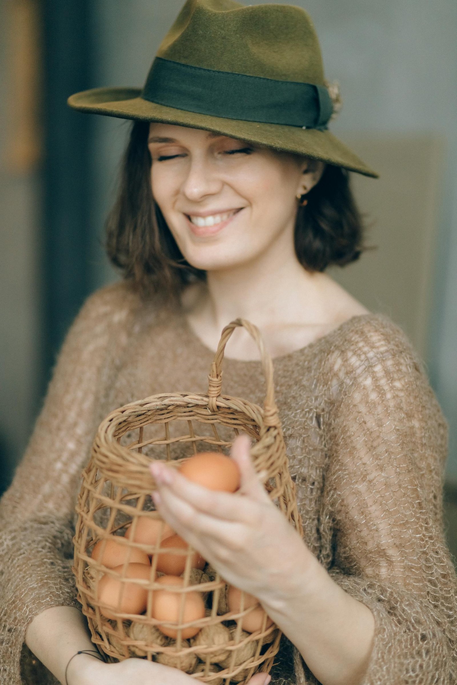 Happy woman in hat holding a wicker basket filled with brown eggs, eyes closed in contentment.